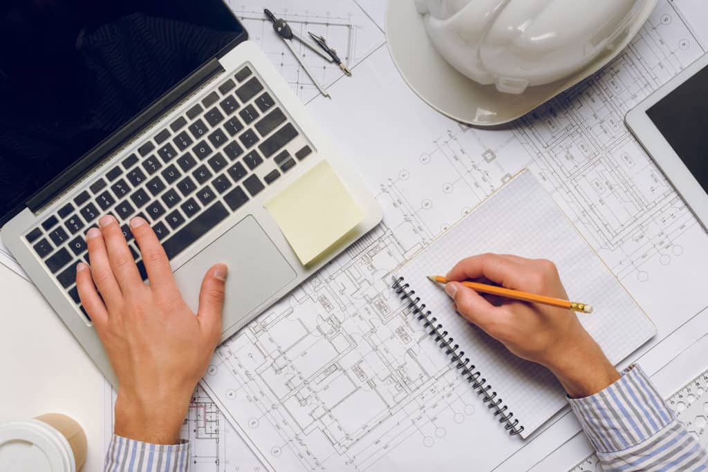 A person works on a laptop and takes notes on an open notebook, surrounded by architectural blueprints, a coffee cup, and a white hard hat—perfecting strategies for home builder marketing.