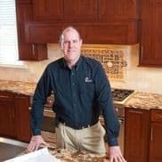 A man stands in a modern kitchen with dark wood cabinets, a patterned backsplash, and a marble countertop, resting his hands on the counter.