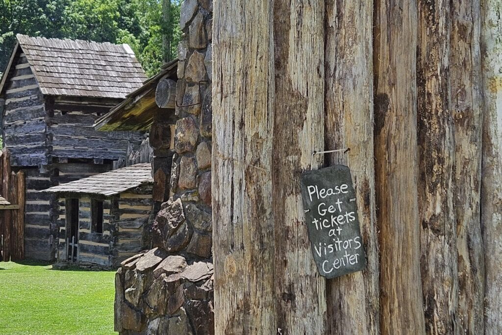 Sign reading “Please Get tickets at Visitors Center” hangs on a wooden wall; rustic log buildings and green grass are visible in the background.