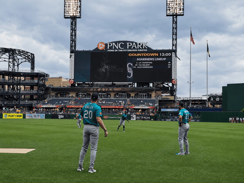 Three baseball players in teal uniforms warm up on the field at PNC Park, with a large scoreboard and stadium lights in the background under a cloudy sky.