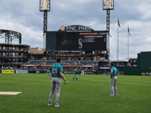 Three baseball players in teal uniforms warm up on the field at PNC Park, with a large scoreboard and stadium lights in the background under a cloudy sky.