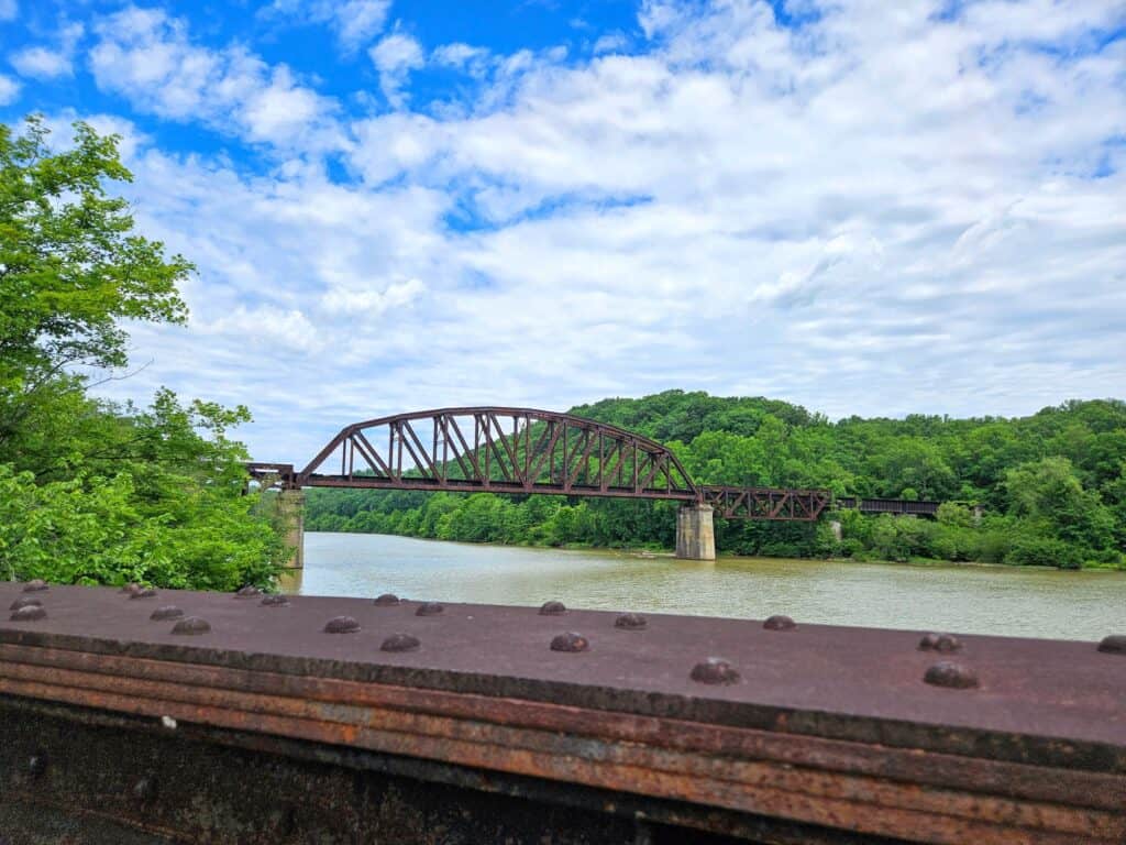 A rusty steel truss bridge spans a wide river, surrounded by green trees and under a partly cloudy blue sky; the photo is taken from behind a metal railing.