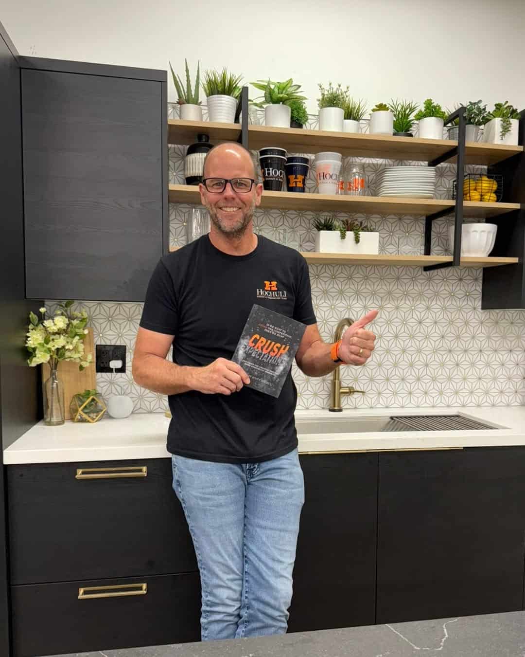 A man stands in a modern kitchen holding a book and giving a thumbs up. Shelves with dishes and potted plants are mounted on the wall behind him.