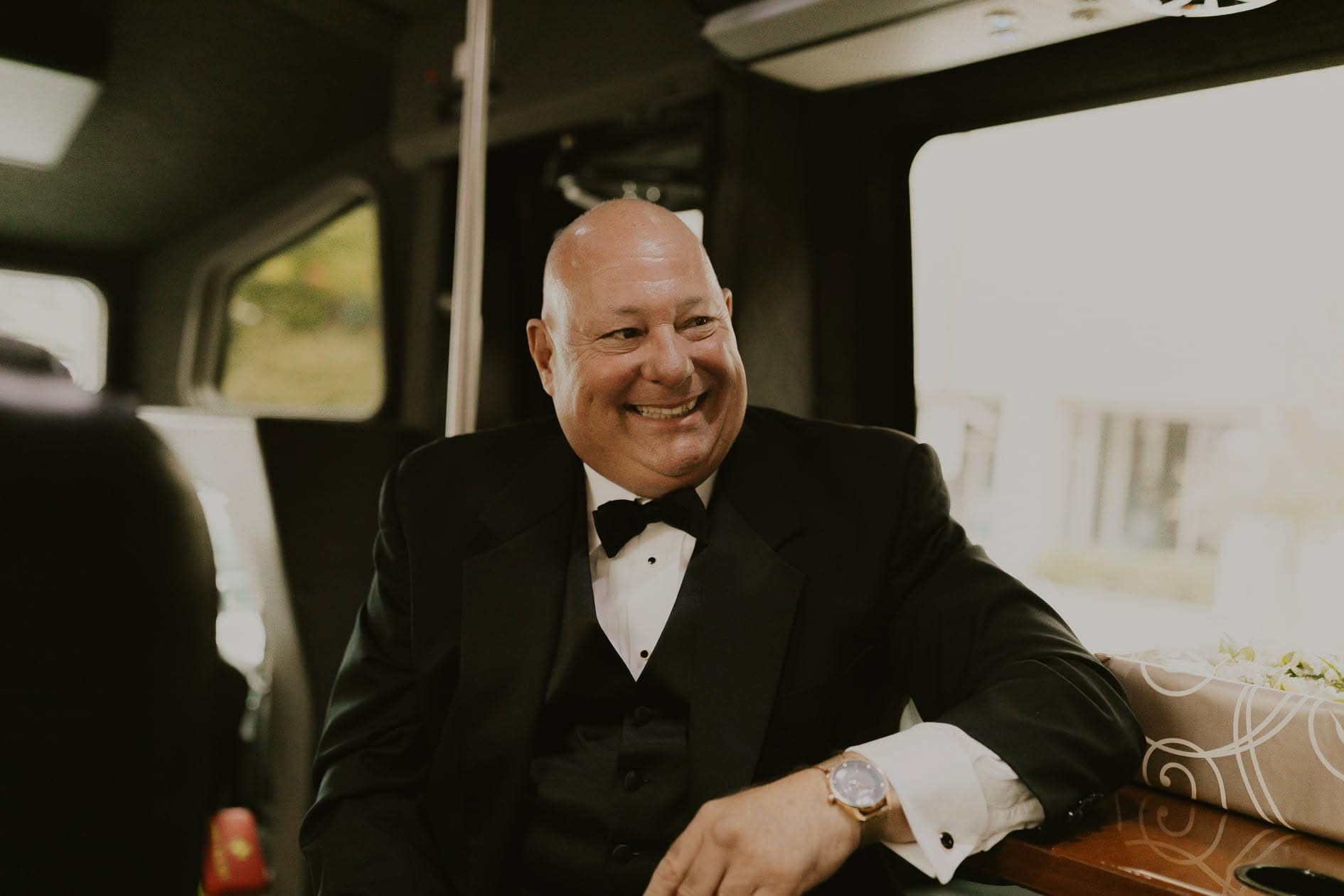 Bald man in a black tuxedo sits and smiles inside a vehicle, resting his arm on the seat next to him.
